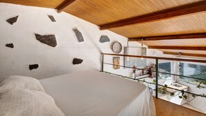 A bedroom with a bed, white walls with stone accents, wooden ceiling beams, a window with a view, and a small kitchenette. - Holiday Home "Tajaraste 2" with Sea & Mountain Views, Private Terraces & Wi-Fi (Hermigua)
