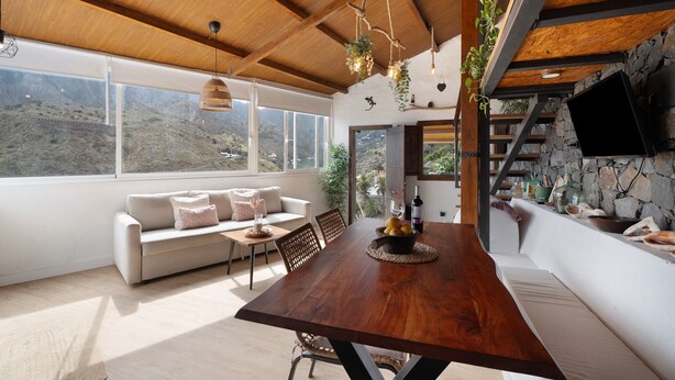 A dining area with a wooden table, white sofa, and a stone wall with a mounted TV. - Holiday Home "Tajaraste 2" with Sea & Mountain Views, Private Terraces & Wi-Fi (Hermigua)