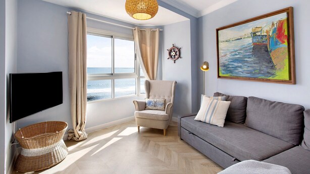 A living room with a television, a sofa, a chair, a window with curtains, a painting, and a ceiling light. - Vacation Apartment 'Ocean Side Arinaga' with Sea View and Wi-Fi (Arinaga)