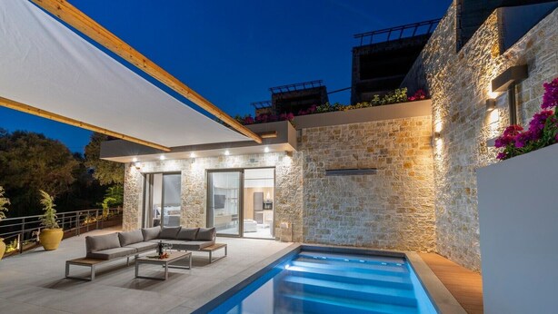 A modern outdoor pool area with a stone wall, a sofa, and a dining table. - Villa "Suite Erifilli" with Sea View, Private Pool & Wi-Fi (Meganisi)