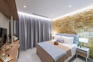 A modern bedroom with a stone wall, a large bed with a grey headboard, and a wooden nightstand with a lamp. - Villa "Suite Diodora" with Private Pool, Terrace & Wi-Fi (Meganisi)