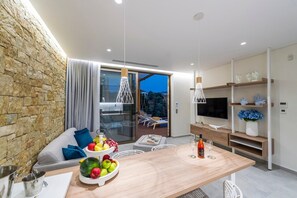 Una sala de estar moderna con una pared de piedra, una zona de comedor con frutas y vino, un televisor y un balcón con mobiliario exterior.