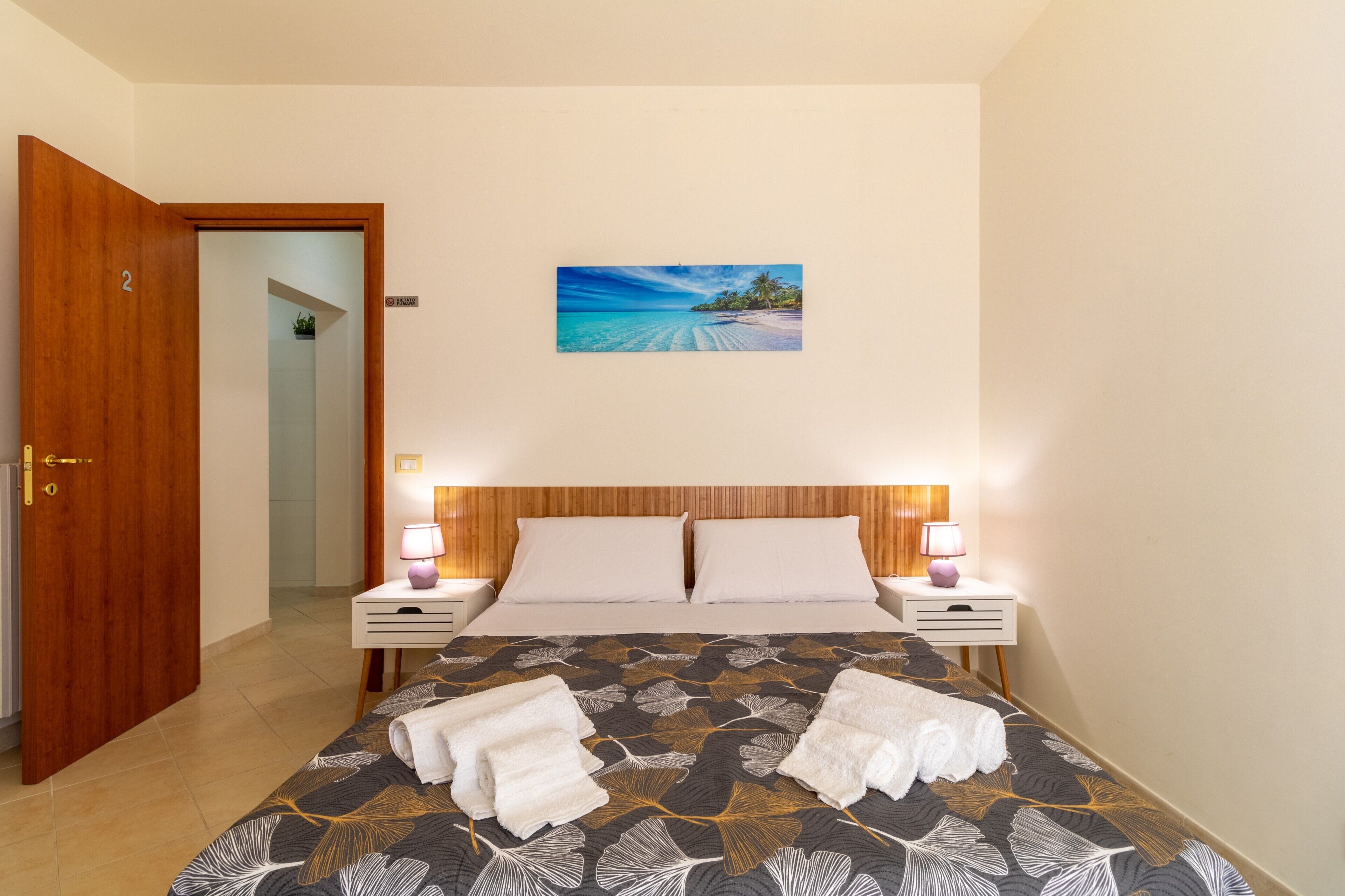 A hotel room with a bed, two bedside tables with lamps, a door, and a painting of a beach scene.