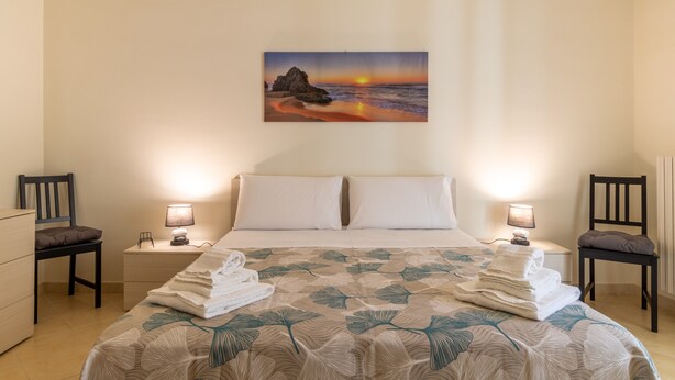 Habitación de hotel con cama, dos mesitas de noche, dos sillas, un cuadro de una puesta de sol y una cama bien hecha con toallas.