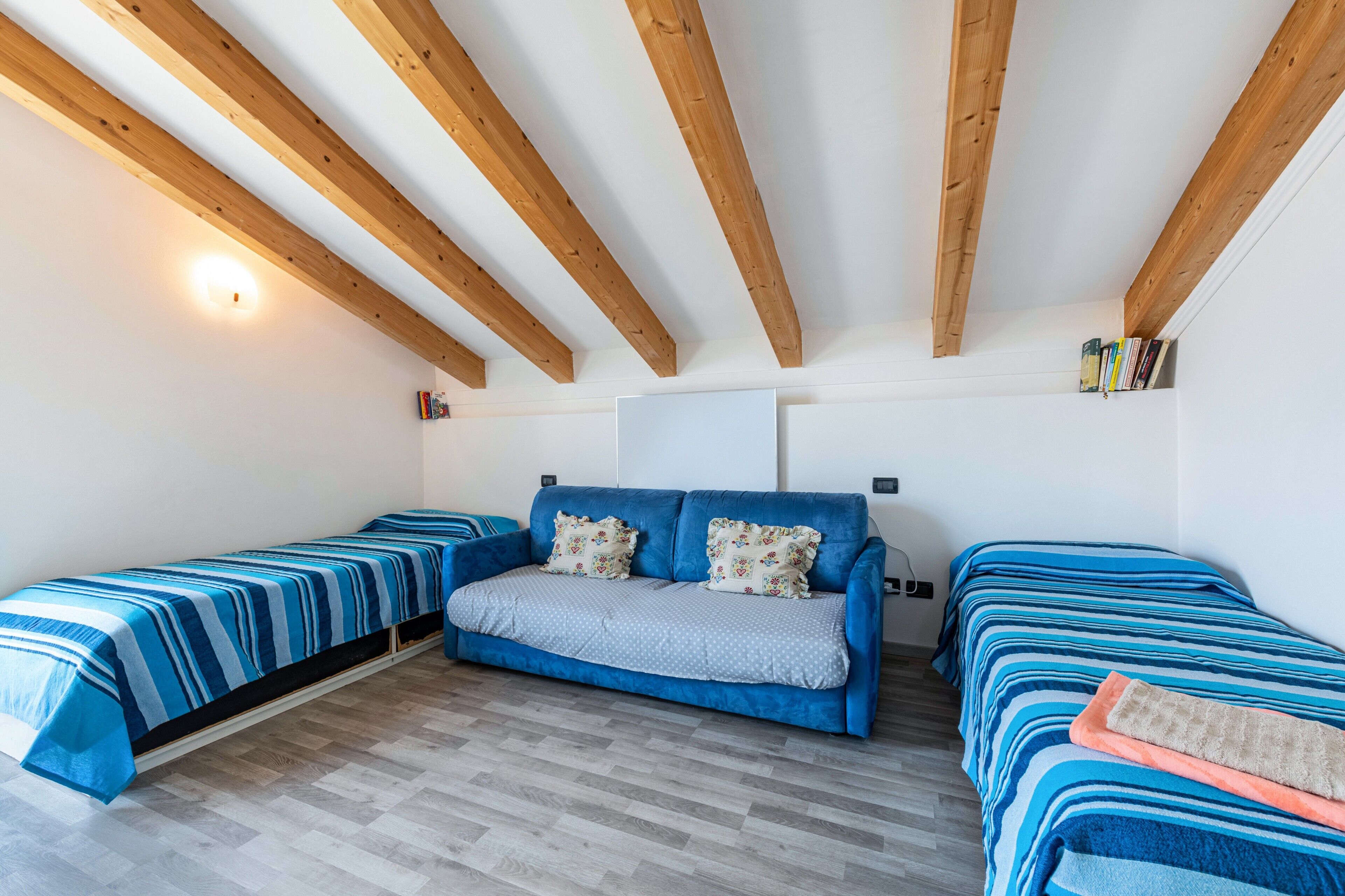 Ein Zimmer mit einem blauen Sofa, zwei Einzelbetten, Holzbalken, einer weißen Deckenleuchte und einem Bücherregal.