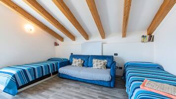 Ein Zimmer mit einem blauen Sofa, zwei Einzelbetten, Holzbalken, einer weißen Deckenleuchte und einem Bücherregal.