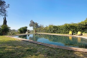 Une piscine rectangulaire bordée de briques, entourée de pelouse et de haies, avec deux chaises jaunes et un hamac à l’arrière-plan.