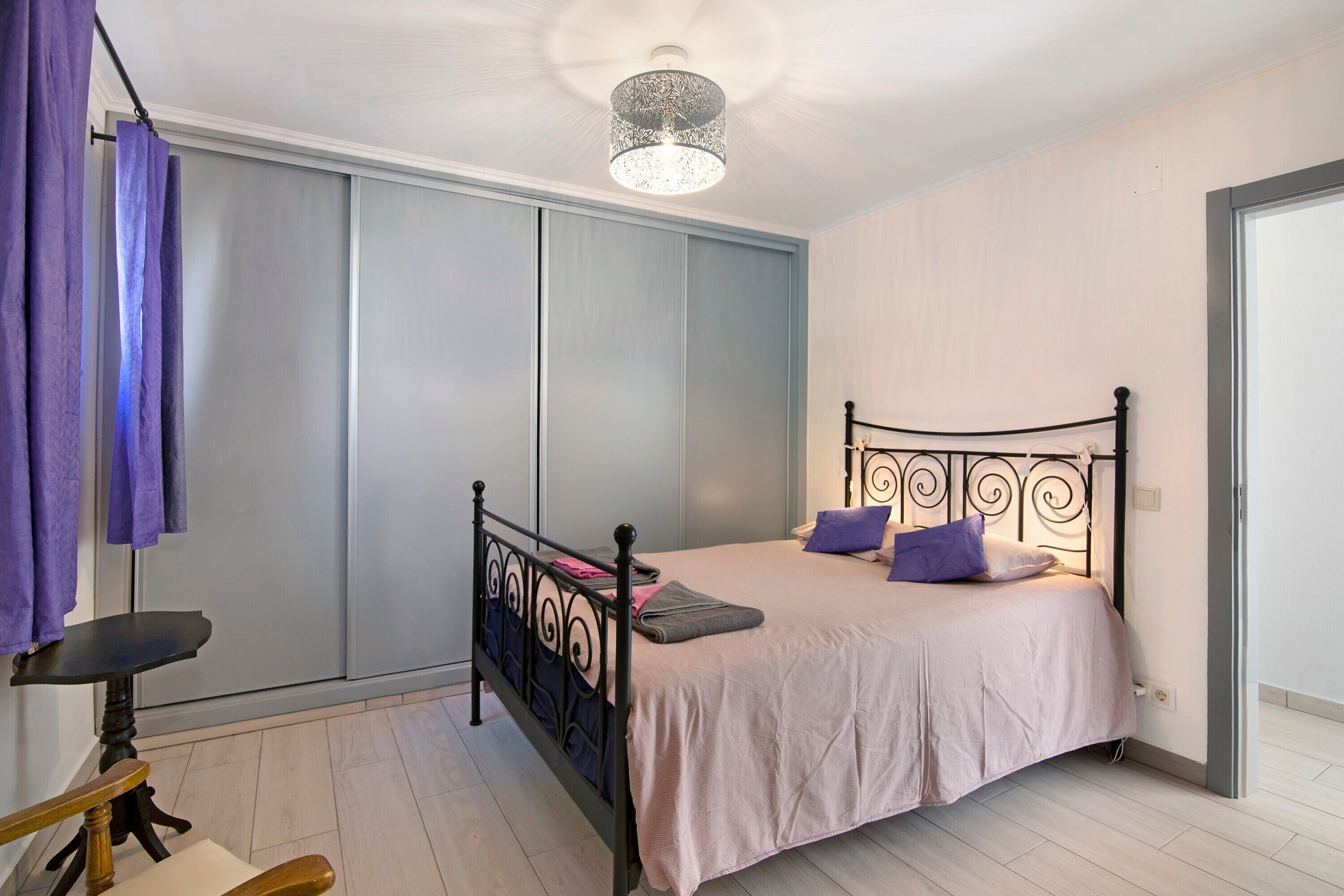 A bedroom with a bed, a table, a chair, a wardrobe, and a ceiling light fixture.