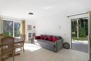 Un salon avec un canapé gris, des coussins rouges, une table à manger avec des chaises, un jeu de fléchettes et une porte-fenêtre coulissante donnant sur un jardin.