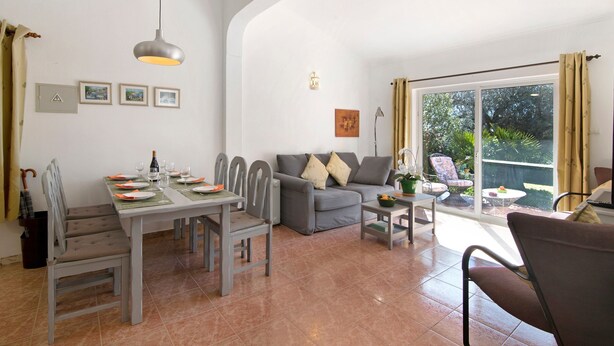 A dining area with a table set for four, a grey sofa, and a patio with a view. - Vacation Home "Arcadia T3" with Private Pool, 2 Terraces & Wi-Fi (São Bartolomeu de Messines)
