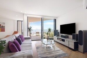 A modern living room with a sofa, TV, speakers, and a balcony with a view. - Apartment "Mare Nostrum" with Sea View, Shared Pool and Wi-Fi (Olhão)