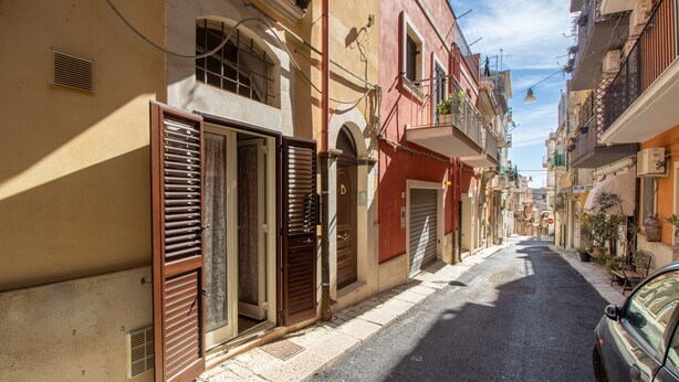Una calle estrecha flanqueada por edificios, algunos con contraventanas, y un coche aparcado a la derecha.