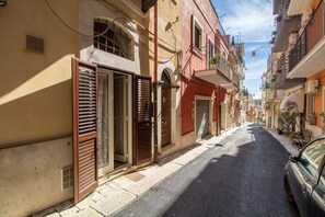 Una stretta via fiancheggiata da edifici, alcuni con persiane, e un'auto parcheggiata a destra.