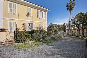 Un bâtiment jaune avec des volets blancs, une allée gravillonnée et un jardin clos.