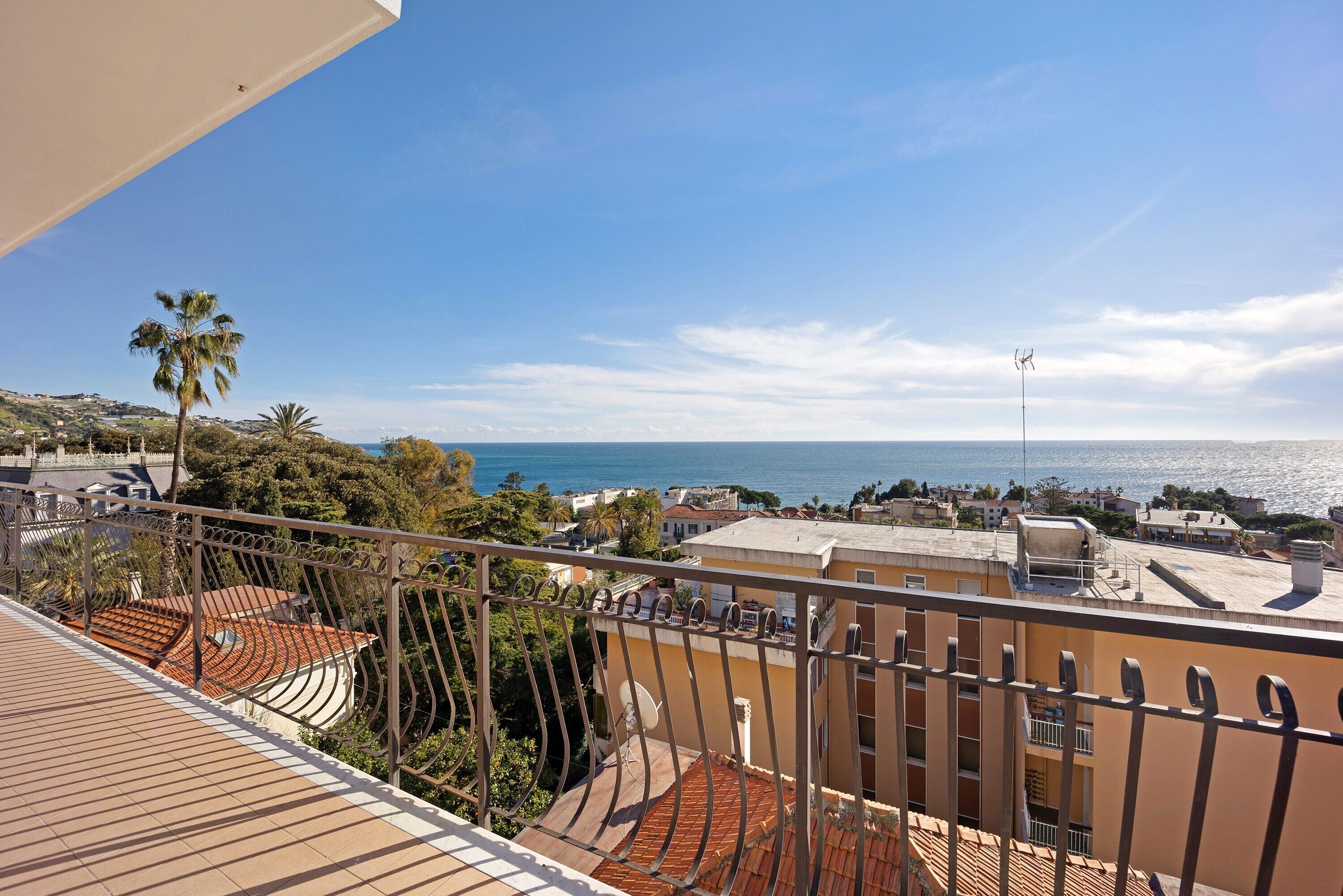 A balcony with a view of the sea, a palm tree, and buildings in the background.