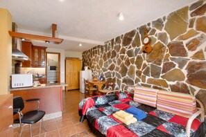 Une petite pièce comprenant un coin cuisine, un lit avec une courtepointe, un mur en pierre et une guitare accrochée à ce mur.