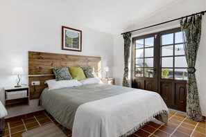 A bedroom with a wooden headboard, a bed with a white bedspread, two bedside lamps, a nightstand, a window with curtains, and a view of the outside.