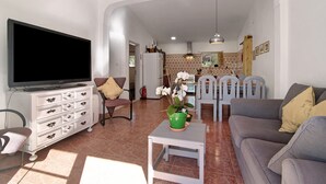 Ett vardagsrum med en tv, en soffa och stolar, ett litet bord med en växt och ett kök i bakgrunden.