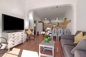 A living room with a TV, sofa, and chairs, a small table with a plant, and a kitchen in the background.
