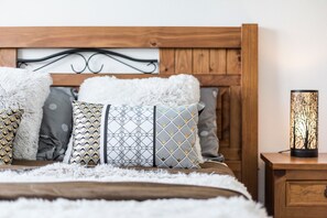 Uma cama com almofadas estampadas, cabeceira de madeira, tapete branco fofo e mesa de cabeceira com luminária decorativa.