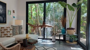 A modern living room with a white sofa, patterned cushions, a large elephant painting, a wooden coffee table, and a view of a lush garden through large glass doors. - Increíble Townhouse con piscina privada (Tulum)