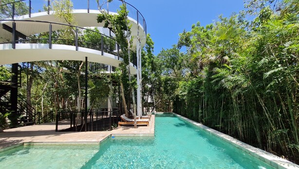 A swimming pool with a clear blue water surface, surrounded by lush greenery and a modern architectural structure with a curved design. - Increíble Townhouse con piscina privada (Tulum)
