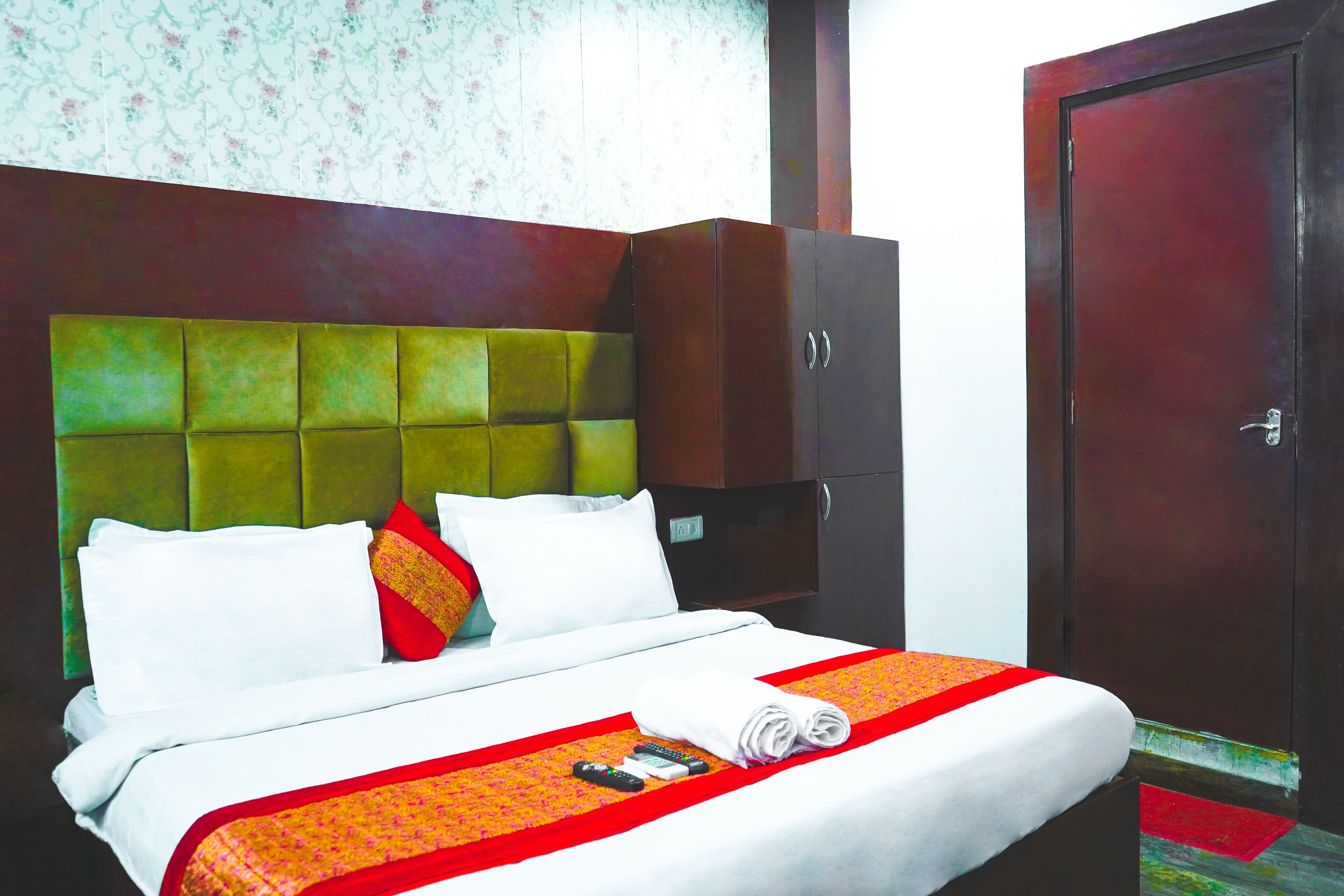 A neatly made bed with a green headboard, white pillows, and a red and orange bedspread. There is a wooden wardrobe beside the bed and a closed door in the background.