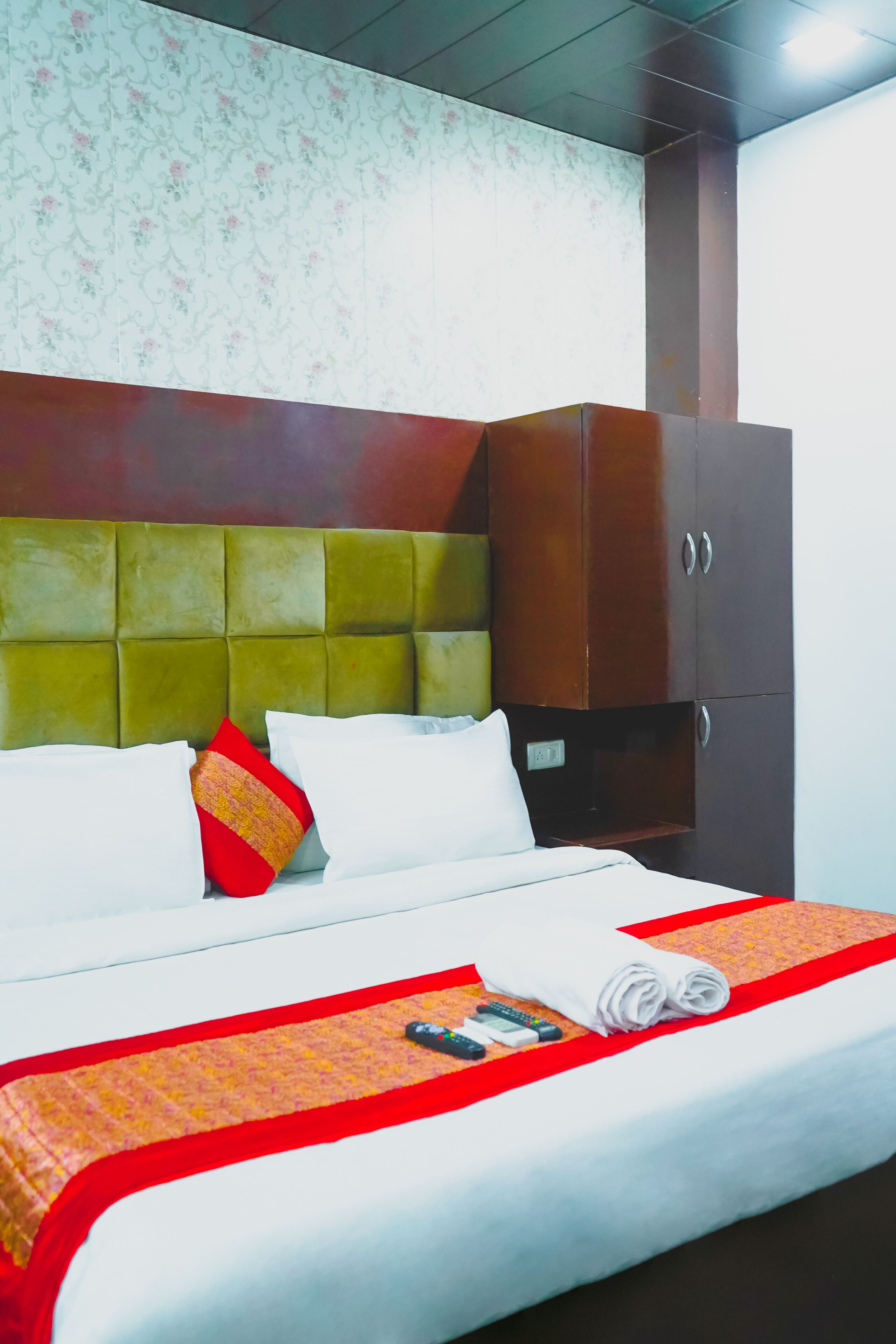A neatly made bed with white linens, a red and gold throw blanket, and a set of remote controls on the bed.