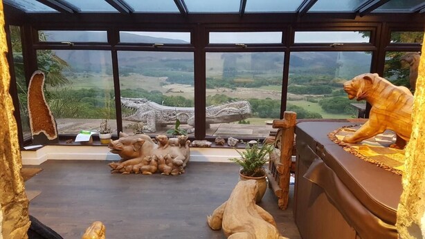 A room with a large glass window offering a view of a mountainous landscape. Inside, there is a wooden desk with a lion statue and several stone sculptures. - Windsor Farm House , Stunning views/hot tub close to Scafell and Wastwater Lake (Wasdale)