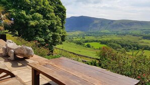 A wooden table with a stone sculpture, overlooking a lush valley with rolling hills and a mountain in the distance. - Windsor Farm House , Stunning views/hot tub close to Scafell and Wastwater Lake (Wasdale)