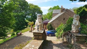 A stone building with two lion statues on the entrance, surrounded by lush greenery and a blue car parked nearby. - Windsor Farm House , Stunning views/hot tub close to Scafell and Wastwater Lake (Wasdale)