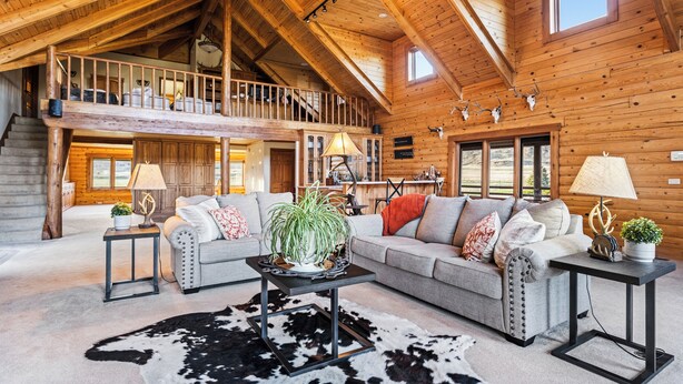 A spacious living area with a high ceiling, a staircase leading to an upper level, and a large window providing natural light. - Sky Creek Ranch Montana  Lodge House
 (Molt)