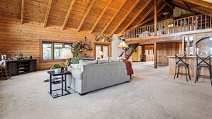 A spacious living area with a high ceiling, wooden beams, a sofa, a coffee table, and a kitchen area with a bar. - Sky Creek Ranch Montana  Lodge House
 (Molt)