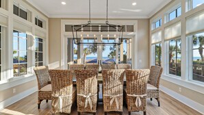 A dining area with wicker chairs and a central chandelier, large windows with views of the beach, and a wooden floor. - New Oceanfront Oasis in Palmetto Dunes with a private pool and EV plug (Hilton Head Island)