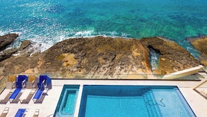 Un área de piscina con agua cristalina, una orilla rocosa y sillas de descanso.
