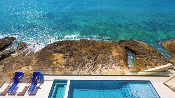 A pool area with clear blue water, a rocky shoreline, and lounge chairs. - SeaDreams 5: Oceanfront Condos w/Pool, Hot Tub, & Rooftop Patio (West Bay)