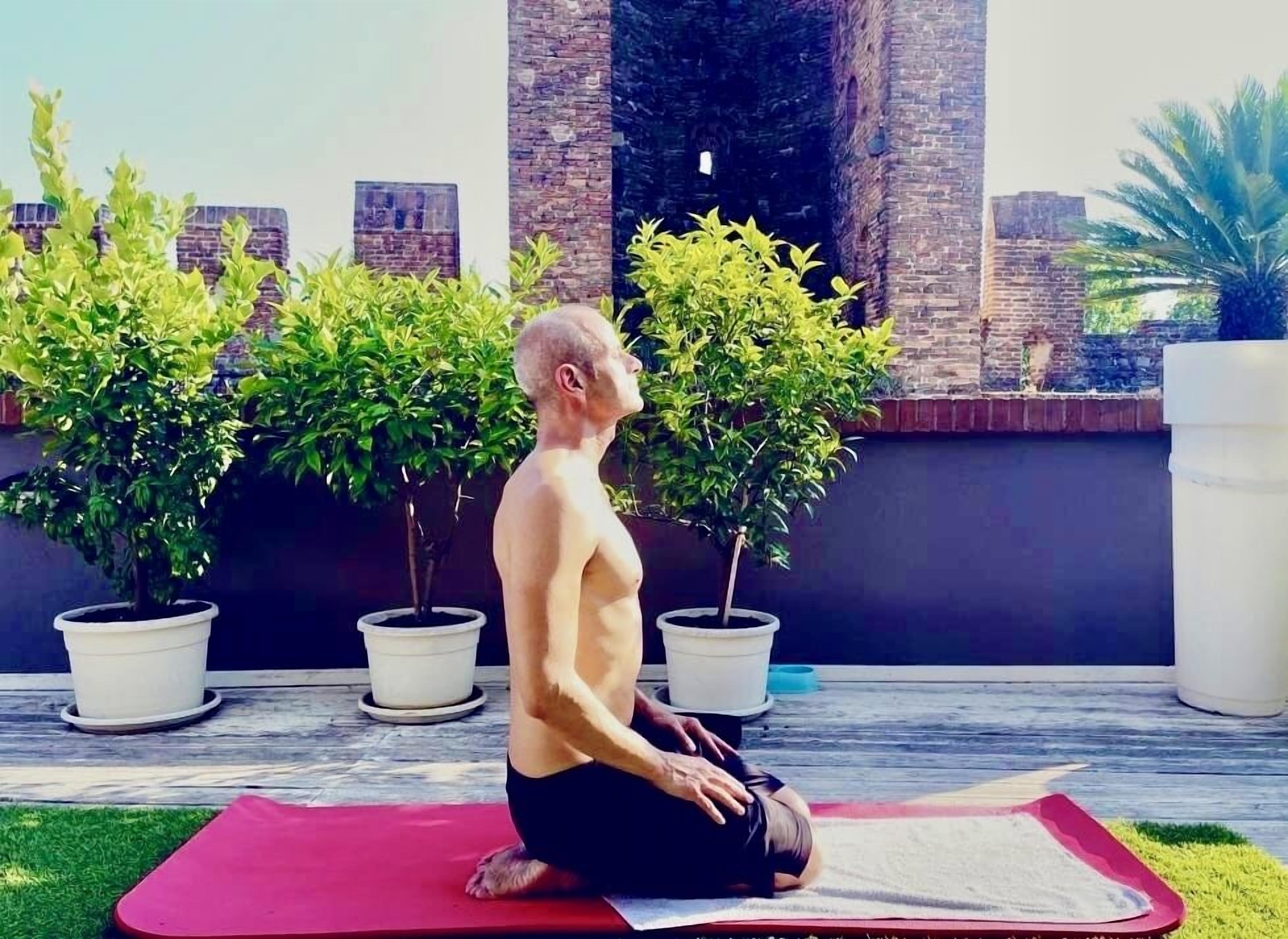 Una persona practicando yoga sobre una esterilla al aire libre, con macetas y un muro de ladrillo de fondo.