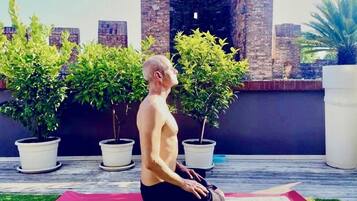 Una persona practicando yoga sobre una esterilla al aire libre, con macetas y un muro de ladrillo de fondo.