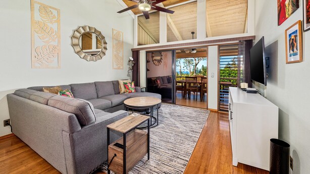 A modern living room with a gray sectional sofa, a round coffee table, and a ceiling fan. There is a TV on the right and a staircase leading to an outdoor area.