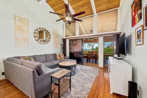 A modern living room with a gray sectional sofa, a round coffee table, and a ceiling fan. There is a TV on the right and a staircase leading to an outdoor area. - Big Island Waikoloa Villas G203 3 Bedroom Condo (Waikoloa)