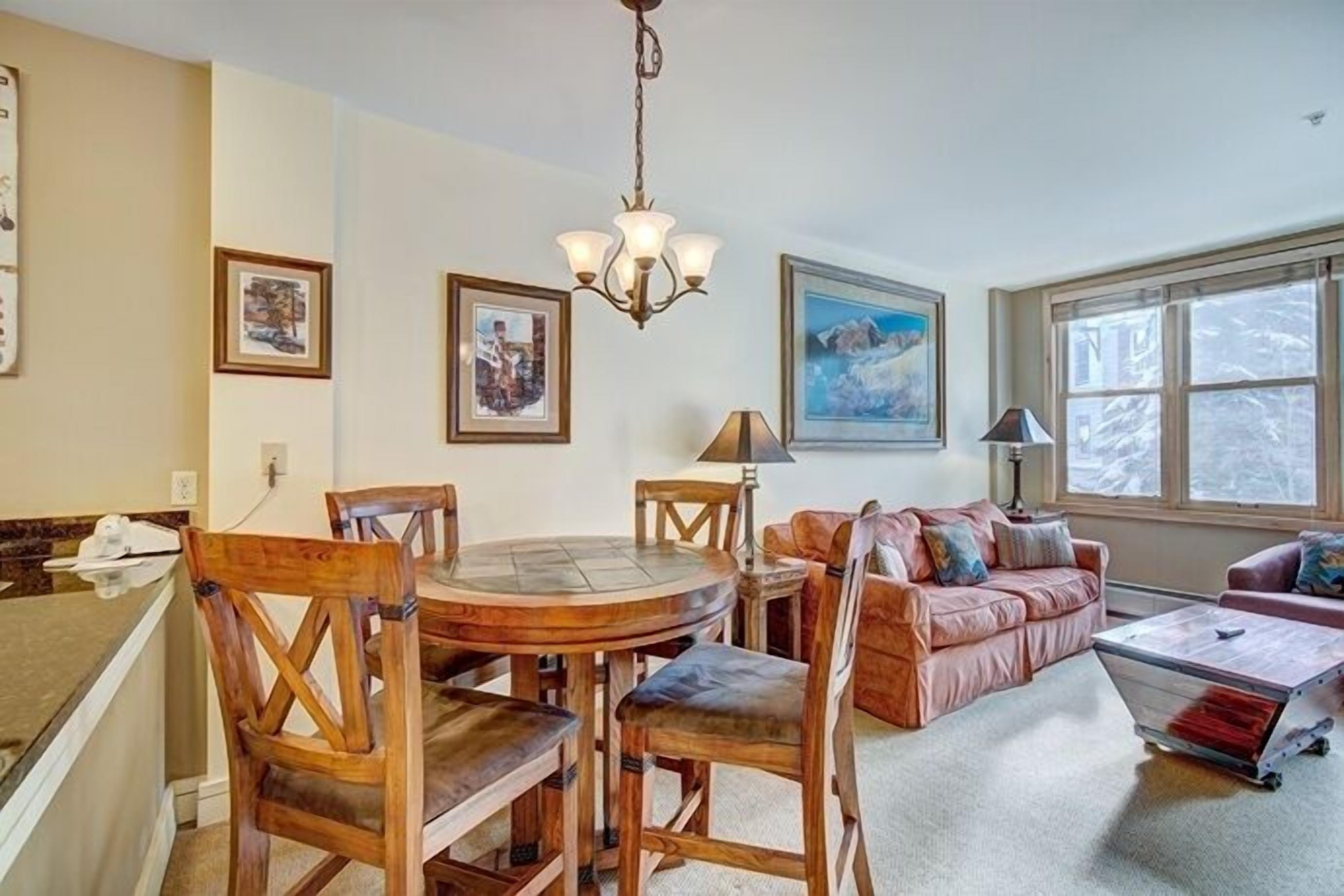 A dining area with a round table and chairs, a chandelier, and a sofa in a living room with a large window.