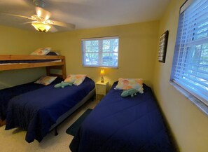 Two single beds with blue bedspreads and matching pillows, a ceiling fan, and a table with a lamp.