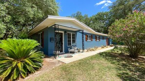 Guests can enjoy a peaceful stay in this charming house with a front porch, surrounded by lush gardens and a large tree.