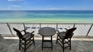 A balcony with two wooden chairs and a table, overlooking a beach with clear turquoise water and a white sandy shore.