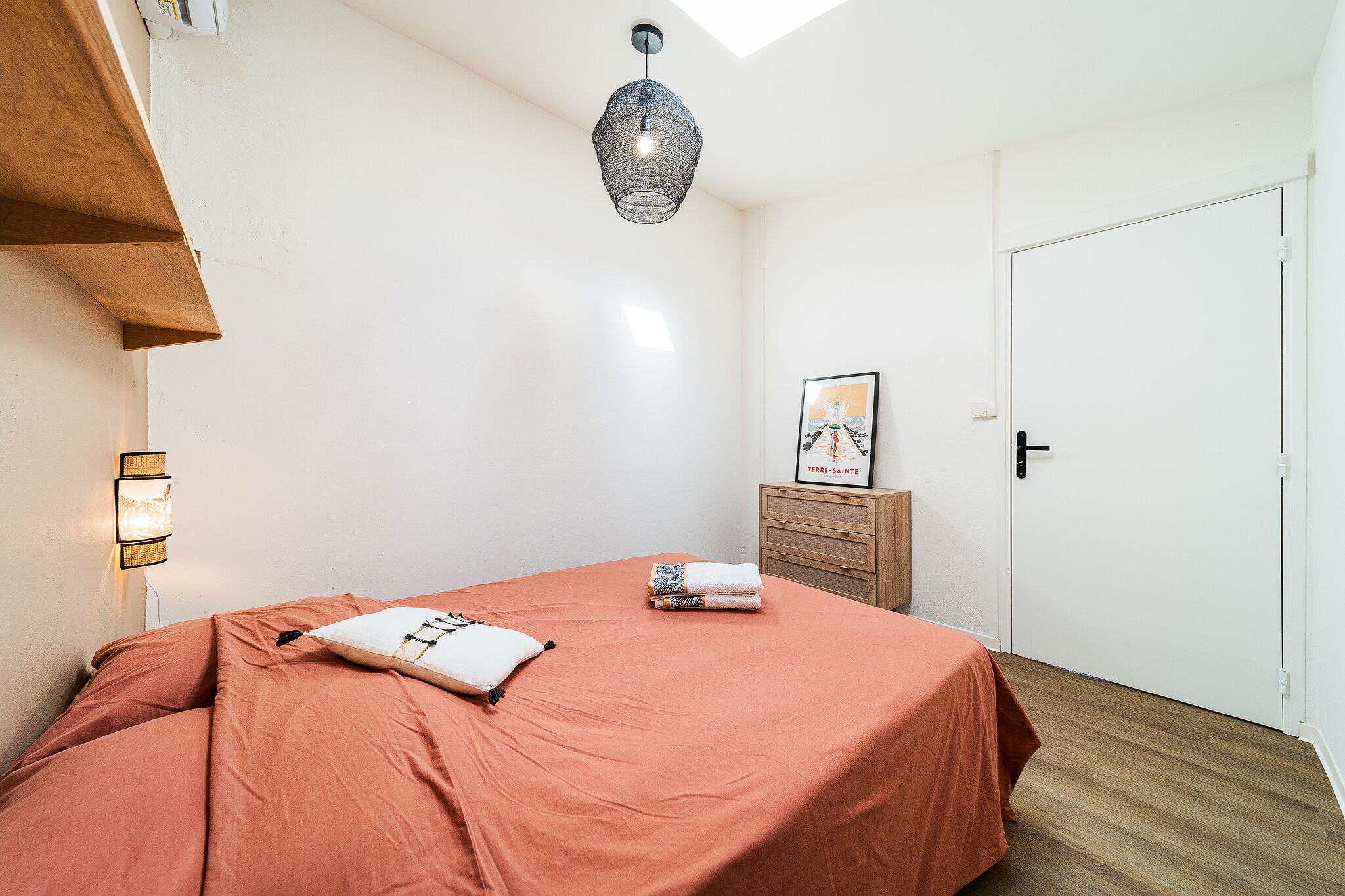 Une chambre à coucher comprenant un lit, une table de chevet, une lampe fixée au mur, un tableau encadré et une porte.