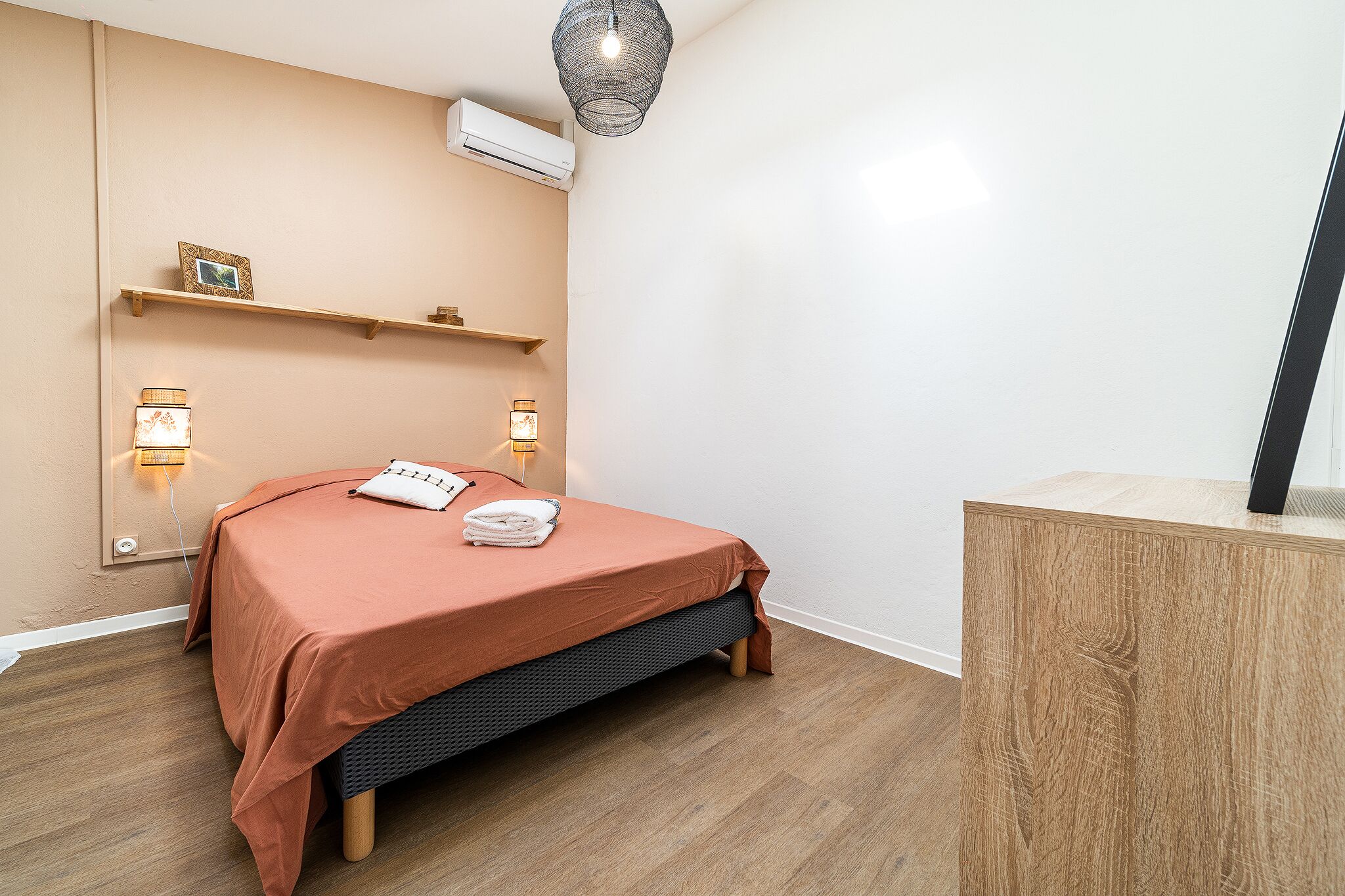 A neatly arranged bedroom with a bed, bedside lamps, a shelf with a picture, and an air conditioning unit.