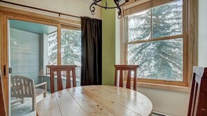 Une table ronde en bois, accompagnée de deux chaises, se trouve dans une pièce dotée de grandes fenêtres offrant une vue sur des arbres couverts de neige.