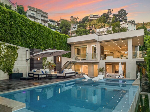Una casa moderna con piscina, mobiliario exterior y un balcón grande con vistas a una zona montañosa.