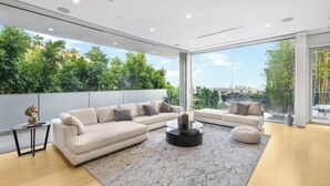 A modern living room with a sectional sofa, a coffee table, and a view of greenery and a cityscape through large windows.