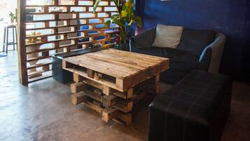 A rustic wooden coffee table with drawers, a black cushioned ottoman, and a blue sofa in a room with a concrete floor and a wooden partition with plants.